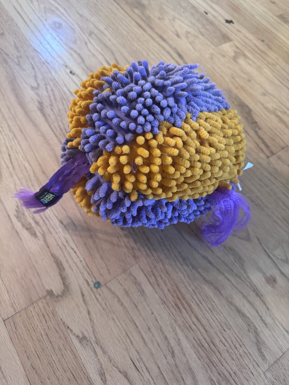 A purple and gold shaggy dog toy on a wood floor after it started ripping apart.